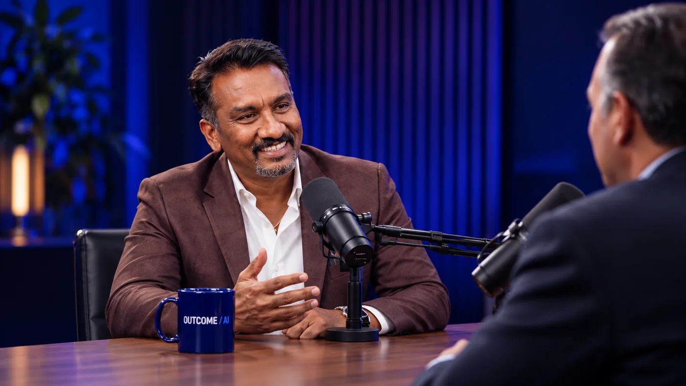 Suresh Devanan in conversation across a studio table, an OUTCOME / AI mug in front of him